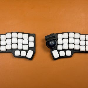 Puede incluir: Un teclado ergonómico dividido que consta de dos unidades base negras con teclas cuadradas blancas. La unidad derecha incluye una bola de seguimiento negra circular grande integrada en la esquina superior izquierda, con una forma contorneada no rectangular.