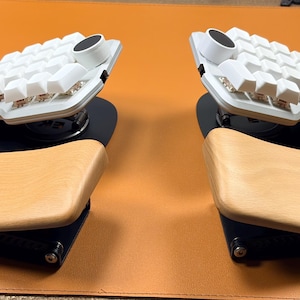 May include: A split ergonomic mechanical keyboard featuring white keycaps, a circular black knob on each half, and light-toned wooden palm rests attached to black metal bases. The keyboard halves are angled and positioned symmetrically.