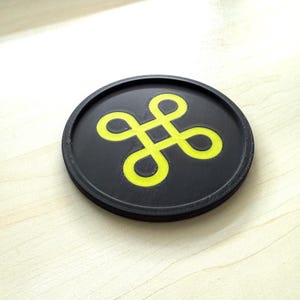 May include: A round, black coaster with a bright yellow, stylized four-leaf clover design in the center. The coaster is made of a textured material and sits on a light-colored wooden surface.