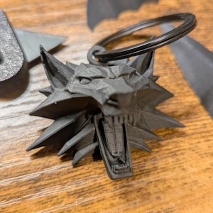 Puede incluir: Un llavero negro impreso en 3D con la cabeza de un lobo con la boca abierta, sujeto a un llavero de metal negro. La cabeza del lobo tiene rasgos afilados y angulosos. Otros objetos grises oscuros impresos en 3D están en el fondo.