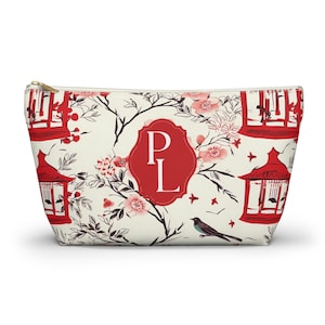 May include: A cream-coloured zippered pouch featuring a repeating pattern of red pagodas, black branches, pink blossoms, and small birds. In the centre, a red scalloped frame contains the white capital letters P and L in a serif font.