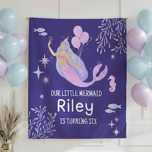 Puede incluir: Una pancarta de cumplea&ntilde;os con tem&aacute;tica de sirena p&uacute;rpura con el texto "Our Little Mermaid Riley is Turning Six". La pancarta presenta una ilustraci&oacute;n de sirena, globos, estrellas y elementos acu&aacute;ticos. Globos de color azul pastel y morado en el fondo.