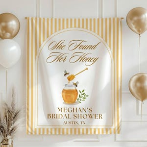 May include: A bridal shower banner with a white background and yellow stripes. The banner features the text "She Found Her Honey" and a graphic of a honey pot with bees. The text "Meghan's Bridal Shower, Austin, TX" is also included.
