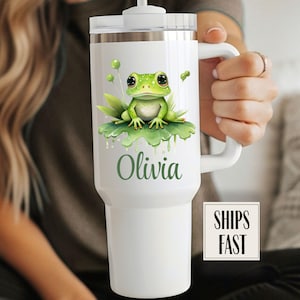 May include: White insulated tumbler featuring a green frog illustration on a lily pad. The name "Olivia" is printed in green script below the frog. The tumbler has a white handle and a white lid with a straw.