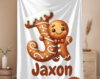 May include: White blanket featuring a gingerbread man holding a gingerbread letter "J" with the name "Jaxon" in brown text. The gingerbread man has a bow tie and is decorated with white icing. A "Ships Fast" sticker is in the bottom right corner.