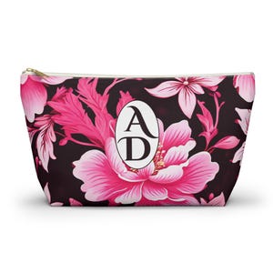Può includere: Pochette per cosmetici con stampa floreale con fiori rosa su sfondo nero. La borsa ha una cerniera dorata e un ovale bianco con le lettere "A" e "D" in nero.