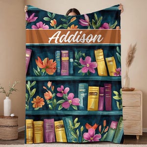 May include: A personalized throw blanket featuring a watercolor design of a bookshelf with colorful books and flowers. The name "Addison" is displayed in white script across a brown band. The blanket has a dark teal background.
