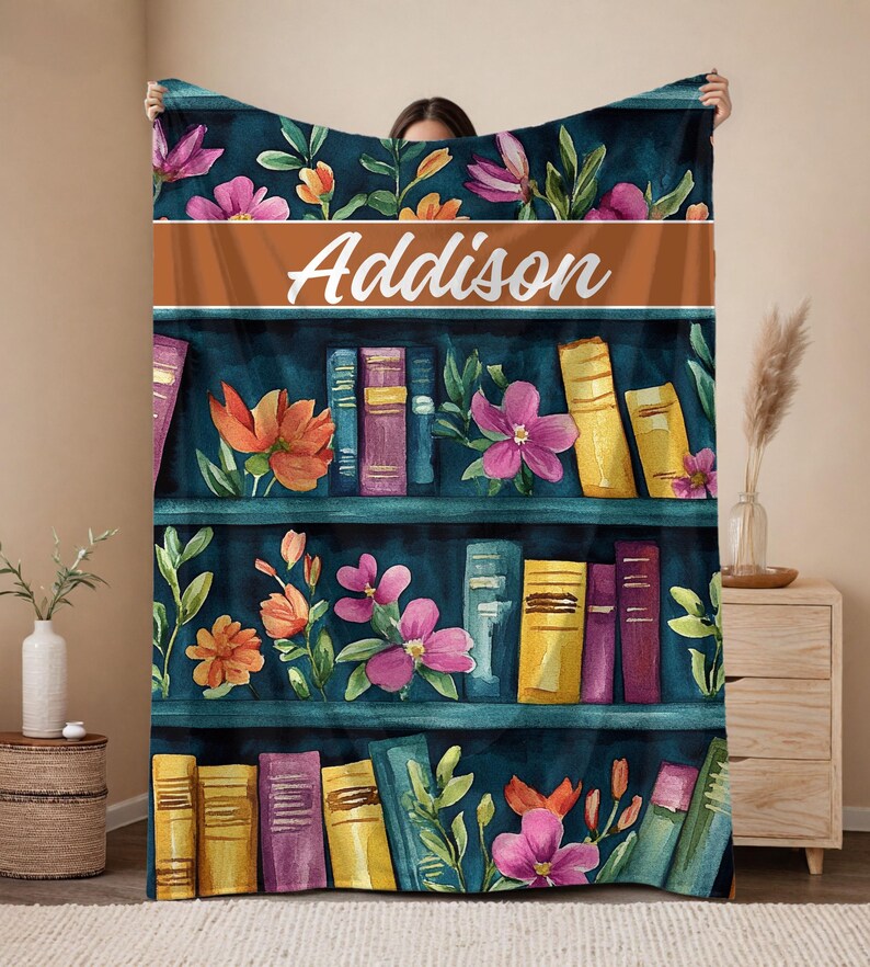 May include: A personalized throw blanket featuring a watercolor design of a bookshelf with colorful books and flowers. The name "Addison" is displayed in white script across a brown band. The blanket has a dark teal background.