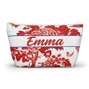 May include: A white cosmetic bag with a red floral pattern and the name "Emma" in red script. The bag has a gold zipper and a blue stripe above and below the name.