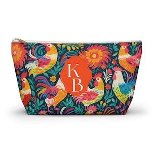 May include: A zipped pouch featuring a dark blue background with a colourful pattern of roosters and flowers. In the centre, an orange scalloped label displays the white capital letters K and B. The pouch has a gold-toned metal zip pull.