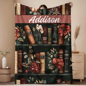 May include: A personalized throw blanket featuring a watercolor design of a bookshelf with books and flowers. The name "Addison" is displayed in white script across a terracotta-colored band. The blanket has a dark green background.