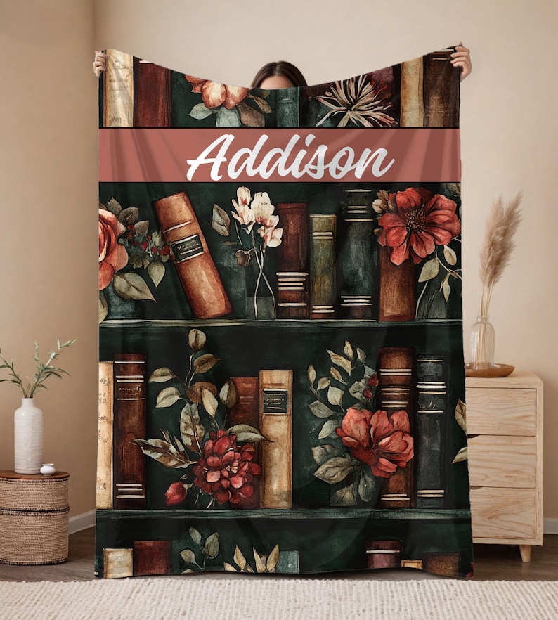 May include: A personalized throw blanket featuring a watercolor design of a bookshelf with books and flowers. The name "Addison" is displayed in white script across a terracotta-colored band. The blanket has a dark green background.