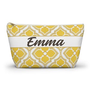 May include: A yellow and white patterned cosmetic bag with a zipper closure. The bag features the name "Emma" in a brown script across a white band. The bag's design includes a repeating floral pattern.