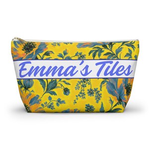 May include: A zipped pouch featuring a vibrant yellow background with a blue floral pattern. A horizontal white band across the centre displays the text Emma's Tiles in a blue script font. The pouch has a gold-toned metal zip pull.
