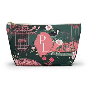 Può includere: Una pochette con cerniera verde scuro caratterizzata da un motivo ripetuto di fiori di ciliegio rosa, un piccolo uccello e una struttura a pagoda stilizzata. Al centro, una cornice smerlata rosa contiene le lettere maiuscole bianche P e L.