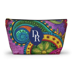 Könnte beinhalten: Kosmetiktasche mit Reißverschluss und lebendigem Paisley-Muster in Lila, Blau, Grün, Gelb und Rot. Die Tasche hat ein marineblaues Emblem mit den Buchstaben "DR". Sie hat einen schwarzen Reißverschluss und einen goldfarbenen Zipper.