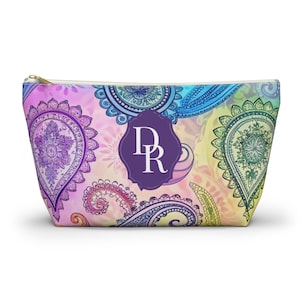May include: A zipped pouch featuring a colourful paisley pattern in shades of purple, blue, yellow, and pink. Centred on the front is a dark purple scalloped emblem containing the white capital letters DR in a serif font.