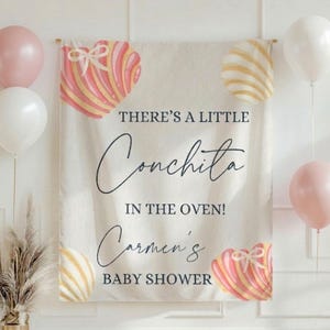 May include: A baby shower banner with the text "THERE'S A LITTLE Conchita IN THE OVEN! Carmen's BABY SHOWER" in black script. The banner has pink and white balloons and decorative heart-shaped designs.