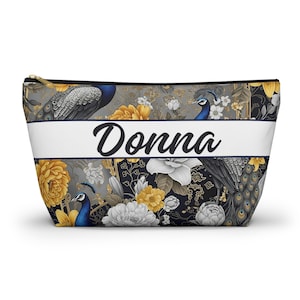 May include: A zipped pouch featuring a repeating pattern of blue peacocks and yellow and white flowers on a dark background. A horizontal white band across the centre displays the name Donna in black script. The pouch has a gold-toned zip.