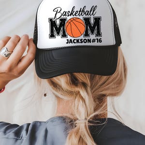 May include: A black and white trucker hat with the text "Basketball MOM" and an orange basketball graphic. The name "JACKSON #16" is printed below the word MOM. The hat has a black brim and mesh back.