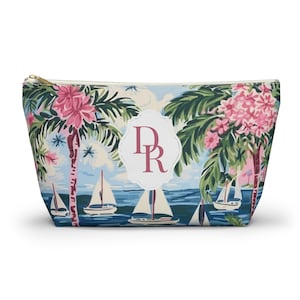 Può includere: Una pochette con cerniera che presenta una scena costiera tropicale con palme, fiori rosa e barche a vela su acqua blu. Un'etichetta bianca smerlata al centro mostra le lettere del monogramma rosa scuro DR. La pochette ha una cerniera dorata.