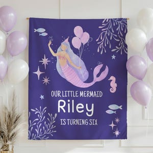 Puede incluir: Una pancarta de cumplea&ntilde;os morada con una ilustraci&oacute;n de sirena. La pancarta dice "OUR LITTLE MERMAID Riley IS TURNING SIX". La sirena es rosa y morada, sosteniendo globos. Globos blancos y morados en el fondo.
