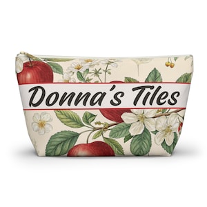 May include: A zipped pouch featuring a cream background with a botanical print of red apples, green leaves, and white blossoms. A horizontal red-bordered white banner across the centre displays the black text Donna's Tiles in a script font.