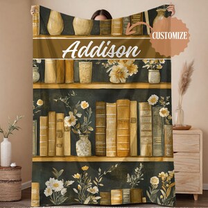 May include: A cozy blanket featuring a bookshelf design with the name "Addison" in white script. The design includes shelves of books, vases, and floral arrangements in shades of gold, brown, and white, set against a dark green background.