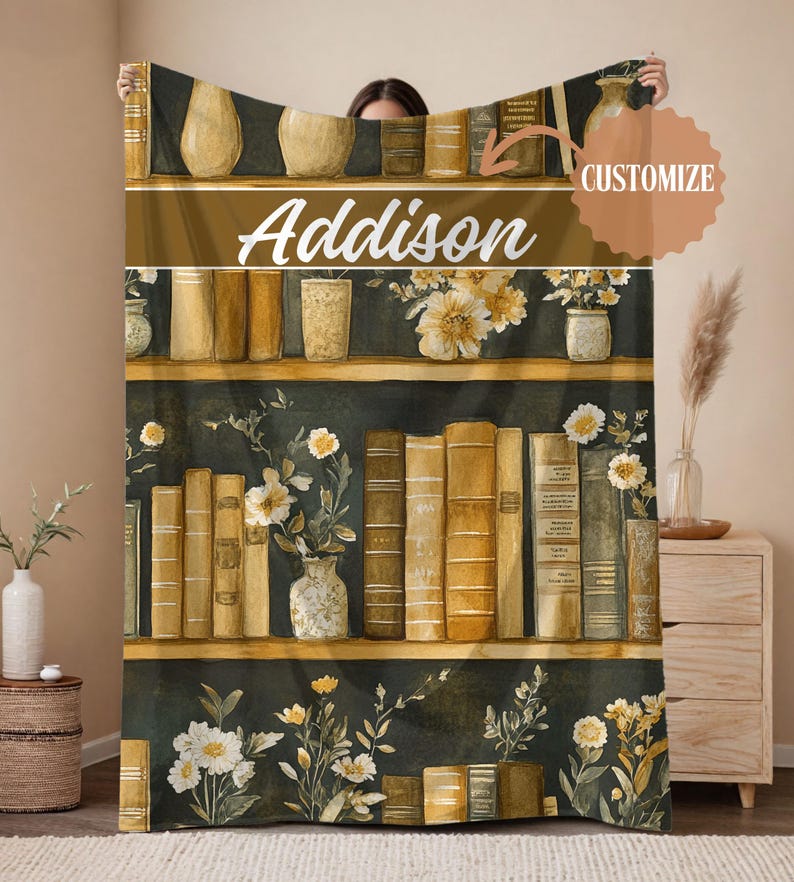 May include: A cozy blanket featuring a bookshelf design with the name "Addison" in white script. The design includes shelves of books, vases, and floral arrangements in shades of gold, brown, and white, set against a dark green background.