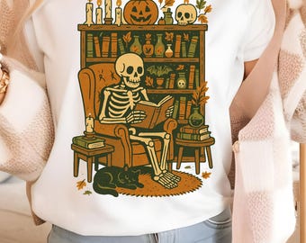 May include: White t-shirt featuring a skeleton reading a book in a cozy armchair, surrounded by Halloween decorations. A black cat rests at the skeleton's feet. The design includes a bookshelf, candles, and a jack-o'-lantern.