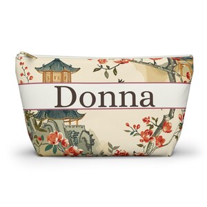 May include: A zipped pouch with a cream background featuring a printed pattern of pagodas, tree branches, and red flowers. A horizontal white band across the centre contains the name Donna in dark brown serif lettering with a thin border.