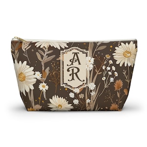 May include: A dark brown zipped pouch featuring a repeating pattern of white and tan daisies and wildflowers. Centered on the front is a rectangular cream-coloured frame containing the stylised black capital letters AR in a serif font.