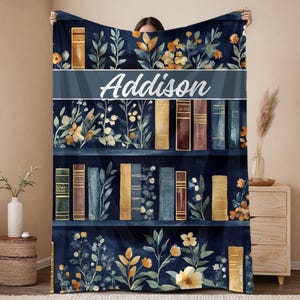 May include: A navy blue blanket featuring a bookshelf design with floral accents and the name "Addison" in white script. The blanket showcases various book spines in shades of brown, gold, and blue, with botanical illustrations.