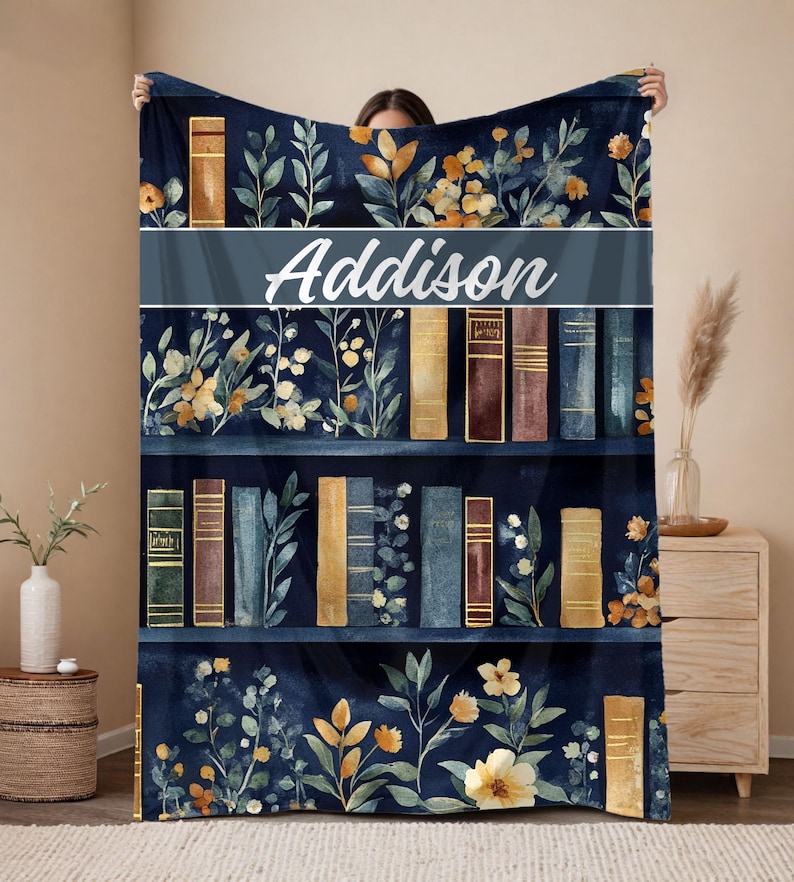 May include: A navy blue blanket featuring a bookshelf design with floral accents and the name "Addison" in white script. The blanket showcases various book spines in shades of brown, gold, and blue, with botanical illustrations.