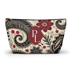 May include: A zipped pouch featuring a cream background with a black, dark red, and grey paisley and floral pattern. A central dark red scalloped label displays the white capital letters PL. The pouch has a black zip closure at the top.