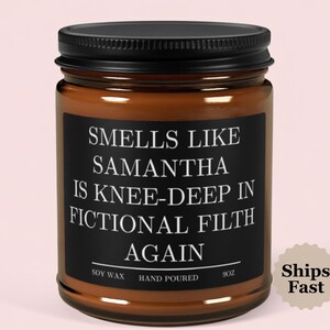 May include: A brown glass jar candle with a black lid. The candle has a black label with white text that reads "Smells Like Samantha Is Knee-Deep In Fictional Filth Again." The label also says "Soy Wax" and "Hand Poured."