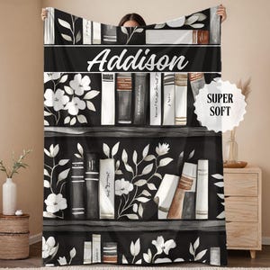 May include: A black and white throw blanket featuring a bookshelf design with floral accents and the name "Addison" in white script. The blanket has a "Super Soft" label.