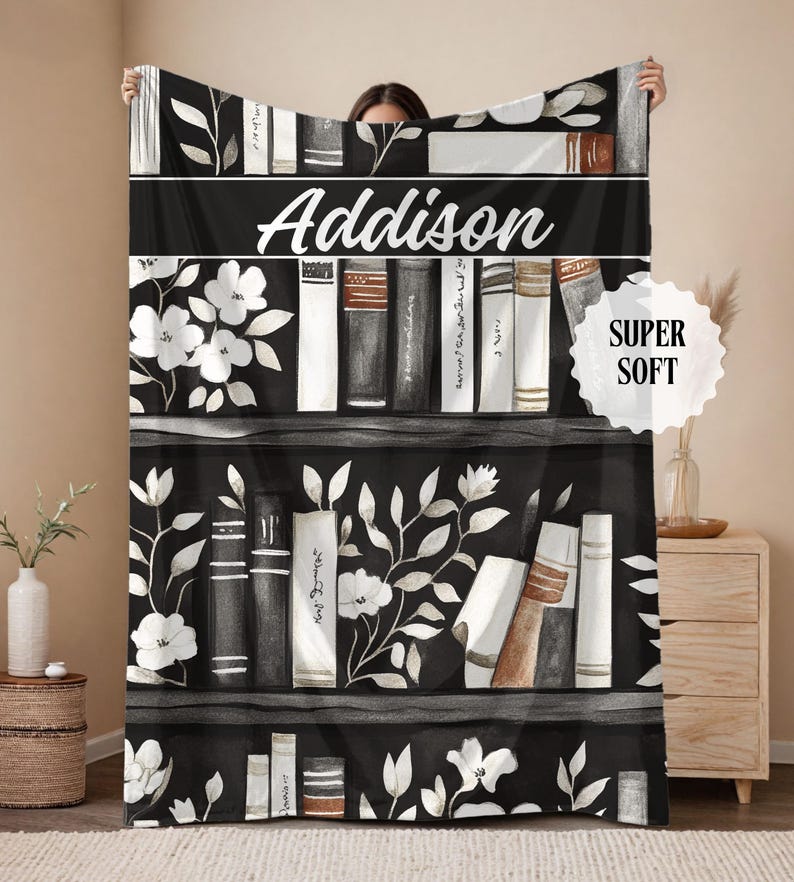 May include: A black and white throw blanket featuring a bookshelf design with floral accents and the name "Addison" in white script. The blanket has a "Super Soft" label.