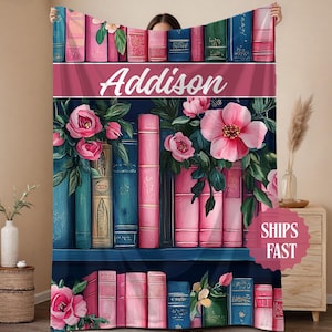 May include: A personalized throw blanket featuring a bookshelf design with pink roses and the name "Addison" in white script. The blanket showcases a navy blue background with colorful books and floral accents.