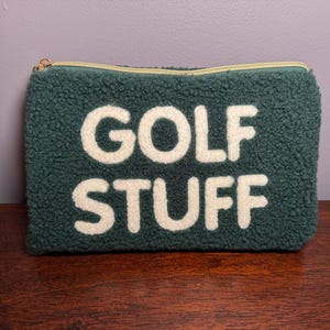 May include: A dark green, textured pouch with the words "GOLF STUFF" in large, white, block letters. The pouch has a gold zipper and is likely used to store golf accessories.