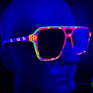 Peut inclure: Lunettes de soleil roses et bleues néon avec un motif floral. Les montures ont un contour vert vif et les verres sont noirs. Les lunettes de soleil sont sur un fond bleu.