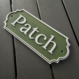 Puede incluir: Un letrero verde y blanco con la palabra "Patch" en letras blancas estilizadas. El letrero tiene un borde decorativo festoneado y dos pequeños agujeros para el montaje. El letrero está sobre una superficie gris oscuro.