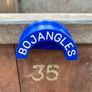 May include: A semi-circular blue plastic sign featuring the white text BOJANGLES in a curved orientation. The object is mounted on a flat surface directly above a metallic house number 35, which is attached to a vertical wooden paneling structure.