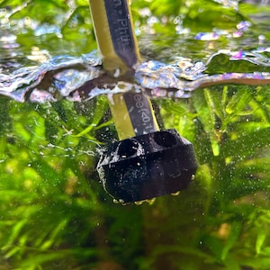 May include: An aquarium scene featuring a black plastic fitting and a yellow and black hose. The hose, marked with "13 mm. Phtha", is submerged in water. Green aquatic plants fill the background, creating a natural setting.