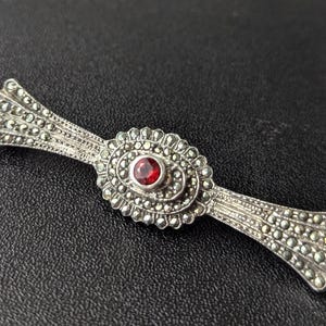 Puede incluir: Broche plateado con un diseño central ovalado con una piedra preciosa roja. El broche tiene una forma simétrica con detalles decorativos y pequeños adornos redondos.