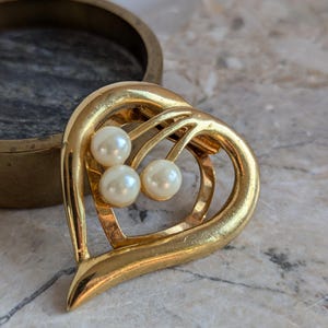 Op de afbeelding: Een gouden broche in de vorm van een hart met drie nepparel accenten.