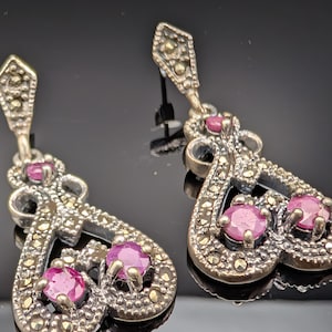 May include: A pair of silver-toned drop earrings with a marcasite and ruby design. Each earring features a decorative top, a central heart-shaped element, and three round, vibrant pink ruby gemstones.