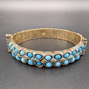 Pode incluir: Uma pulseira de bracelete de latão vintage com duas fileiras de pedras redondas azuis claras. A pulseira tem um fecho e uma aparência antiga e manchada. É uma joia.