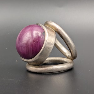 Könnte beinhalten: Ein Silberring mit einem großen, ovalen, tiefvioletten Stein. Der Stein ist in eine silberne Lünette gefasst und von einem silbernen Band umgeben, das sich um den Finger krümmt. Der Ring hat ein modernes, geometrisches Design.