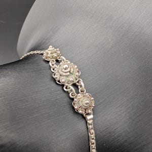 May include: A silver-toned bracelet featuring three ornate, dome-shaped filigree charms connected by small metal links. The bracelet has a flat, woven chain design and displays intricate, textured metalwork on each of the circular focal pieces.